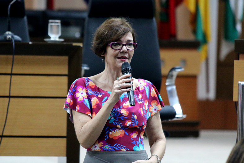 Mulher branca usando óculos e blusa colorida fala em pé ao microfone em um auditório.