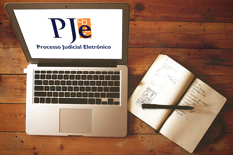 Visão de cima de uma mesa de madeira onde repousa um laptop com a tela aberta projetando a marca do PJe (Processo Judicila Eletrô