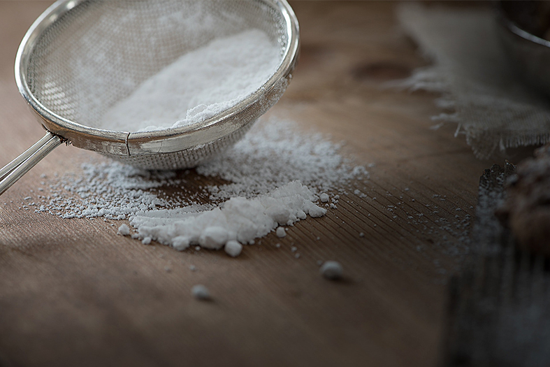 Detalhe de uma quantidade de açúcar branco sendo peneirada sobre mesa de madeira.