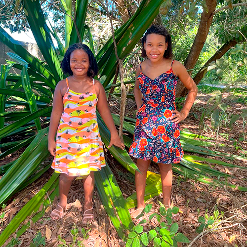 Ana Lúcia de 12 anos e Marlúcia de 8 anos, duas meninas negras de cabelos anelados. Ambas estão vestindo vestidos com motivos florais. Elas estão em um parque arborizado e estão sorrindo para a câmera.