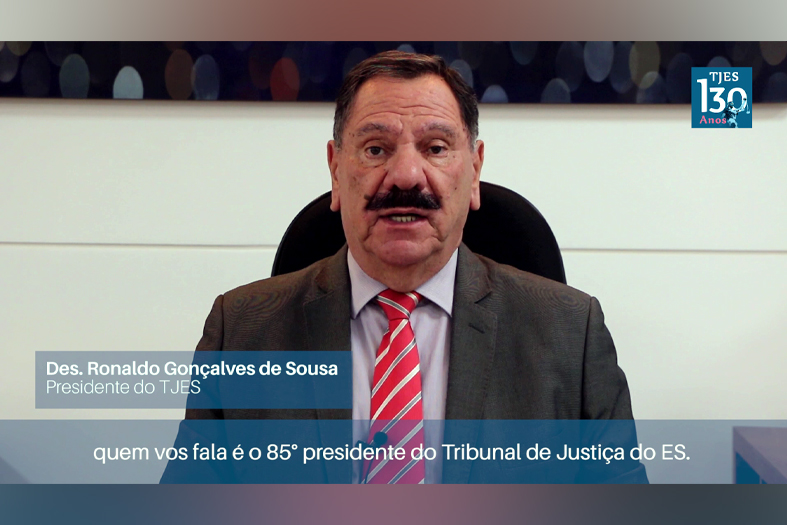 homem de pele branca, com bigode e cabelos escuro e trajando terno ocre e gravata rosa fala para câmera.