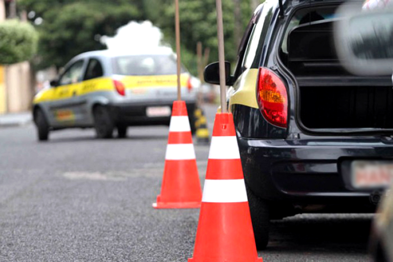 Carros de uma auto escola. Em primeiro plano um veículo de cor preta fazendo baliza. No plano de fundo um veículo cinza manobrando.