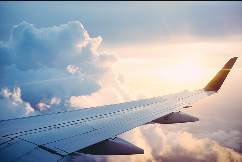 Detalhe da asa de um avião, ao fundo um céu azul com muitas nuvens ensolaradas.