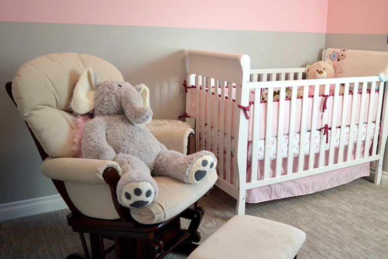 Quarto de recém nascido com decoração em tons da cor rosa, no berço vemos um urso de pelúcia bege e na cadeira de balanço um elefante de pelúcia na cor cinza claro.
