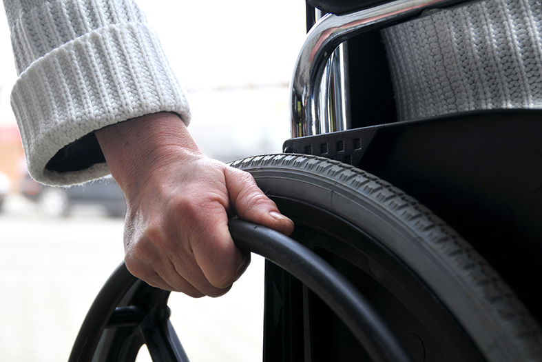 Detalhe de uma pessoa com vestido xadrez sentada em uma cadeira de rodas onde a vemos de costas com a mão segurando a alavanca da roda.