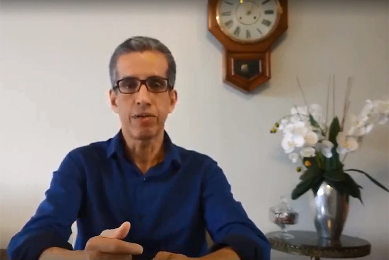 O coordenador do Programa Justiça Presente do Estado do Espírito Santo, Carlos Pinheiro, falando à câmera filmadora. Ao fundo um vaso de flores brancas e preso à parede um relógio de ponteiro.