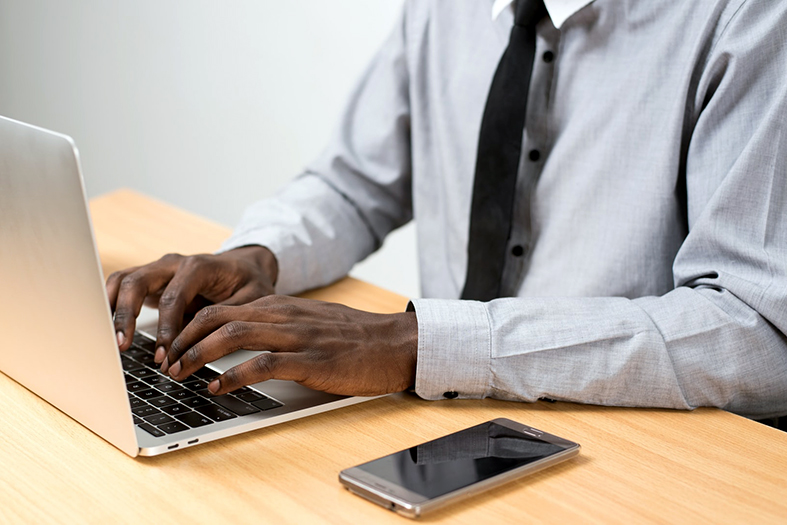 Pessoa negra vestindo uma camiseta social na cor cinza e uma gravata na cor preta digita em um laptop. ao seu lado esquerdo, sobre a mesa de madeira, repousa um smartphone.