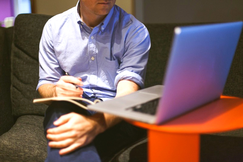 homem faz anotações em um caderno em seu colo enquanto olha para um laptop