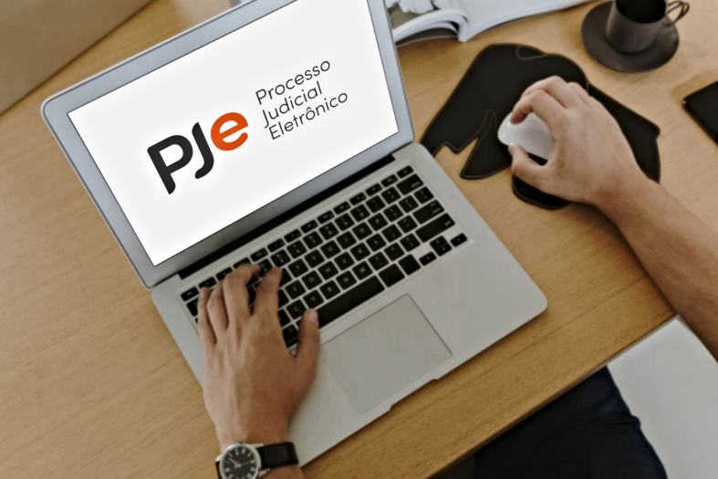 Fotografia dos braços de uma pessoa utilizando um laptop. Na tela, sobre um fundo branco, está projetado o logotipo do Pje e ao lado dele o texto "Processo Judicial Eletrônico".