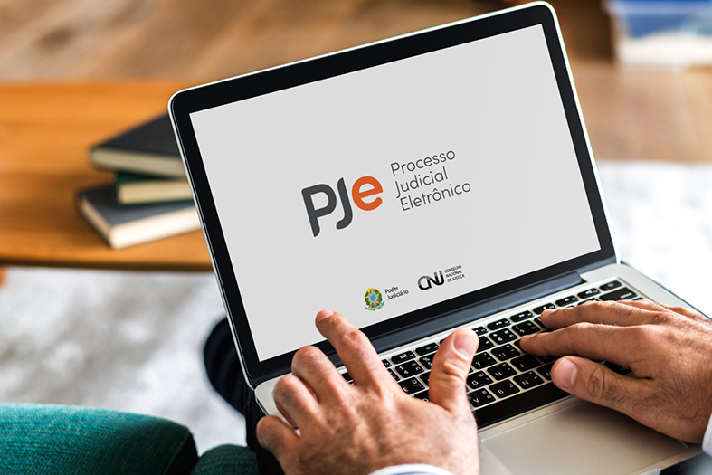 Fotografia dos braços de uma pessoa utilizando um laptop. Na tela, sobre um fundo branco, está projetado o logotipo do Pje e ao lado dele o texto "Processo Judicial Eletrônico".
