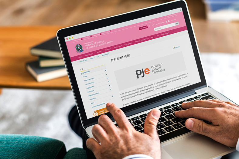 detalhe das mãos de uma pessoa utilizando um laptop. Na tela aparece uma a página do Pje no site do TJES, no topo da página o logotipo do Pje e ao lado dele o texto "Processo Judicial Eletrônico".