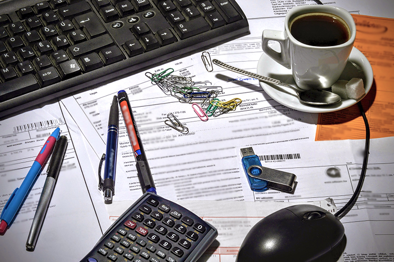 Mesa cheia de contas em papel, uma xícara cheia de café, um teclado de computador, um mouse, quatro canetas e vários clipes de papel.