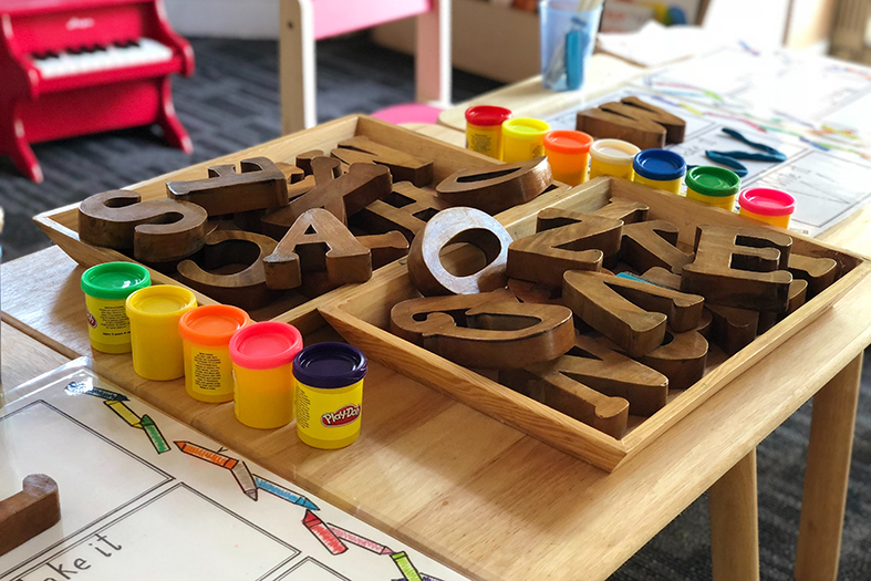 Em uma creche, uma mesa de madeira com várias letras de madeira mais escura e potes de tinta.