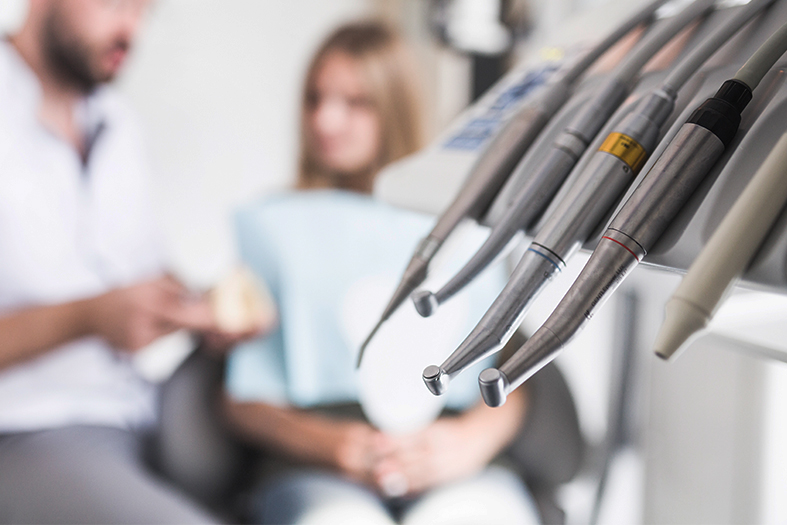 Detalhe de instrumentos de ortodontia em primeiro plano e em segundo, mais desfocado, duas figuras: um dentista e sua paciente.