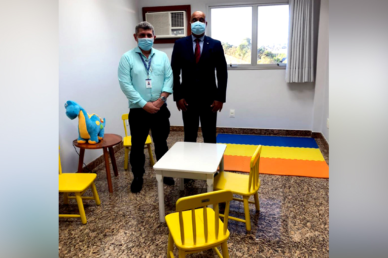 Dois homens, um de pele negra e estatura alta e o outro de branca e estatura mediana posam para foto. em volta deles vários móveis e objetos adequados para crianças brincarem.