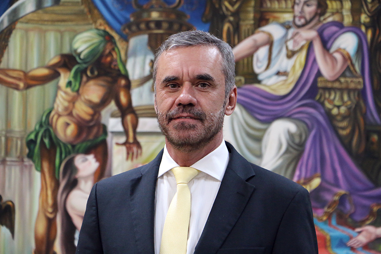 desembargador fernando bravin posando para uma foto com terno na com azul escuro e gravata amarelo claro. Ao fundo desfocado uma pintura.