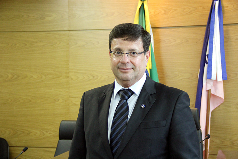 Des. Fernando Zardini posando para uma fotografia. Ele está de pé, ao fundo e atrás dele estão as bandeiras do brasil e a do estado do espírito santo.