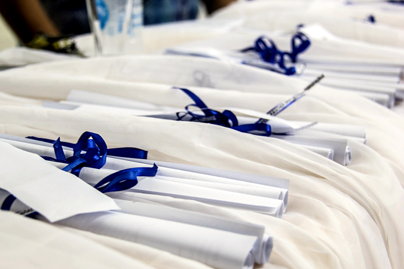 Diplomas enrolados como canudos dispostos sobre mesa com toalha branca.