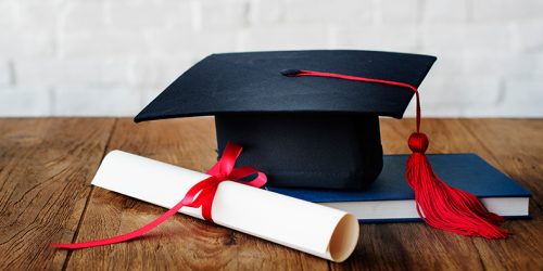 Diploma, chapéu de formando e livro repousam sobre mesa de madeira.