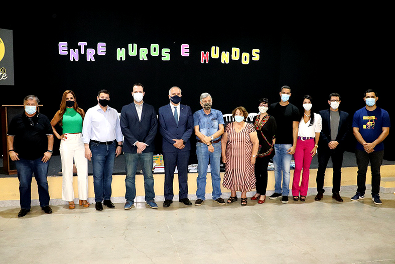 Grupo de onze pessoas, entre homens e mulheres adultos, posam para foto em uma espaço amplo. Atrás do grupo, em letras coloridas, lê-se: entre muros e mundos.