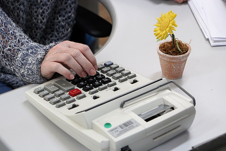 Detalhe de mão feminina digitando em uma calculadora especializada para cálculos financeiros. Decorando a mesa vemos uma flor amarela.