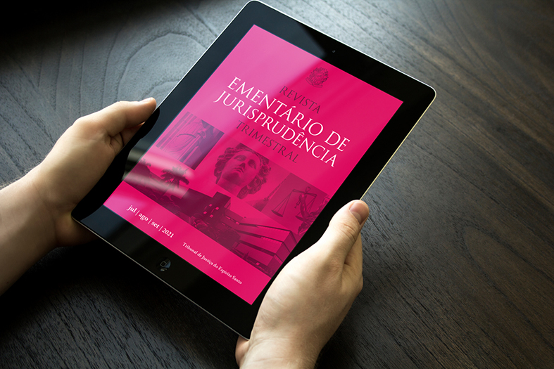 Detalhe de mãos segurando um tablet que exibe em sua tela a capa da última edição da revista ementário trimestral, de cor rosa.