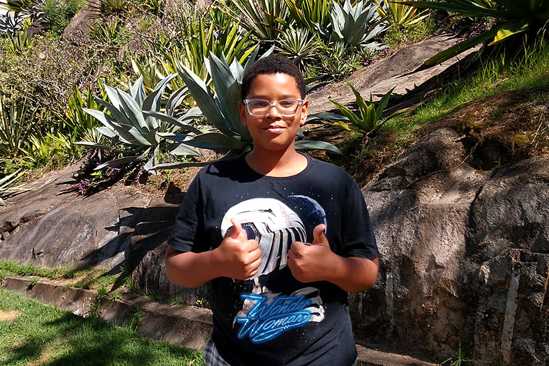 menino de pele negra vestindo uma camiseta estampada e fazendo sinal de "joia" com as duas mãos. Ao fundo um parque arborizado.