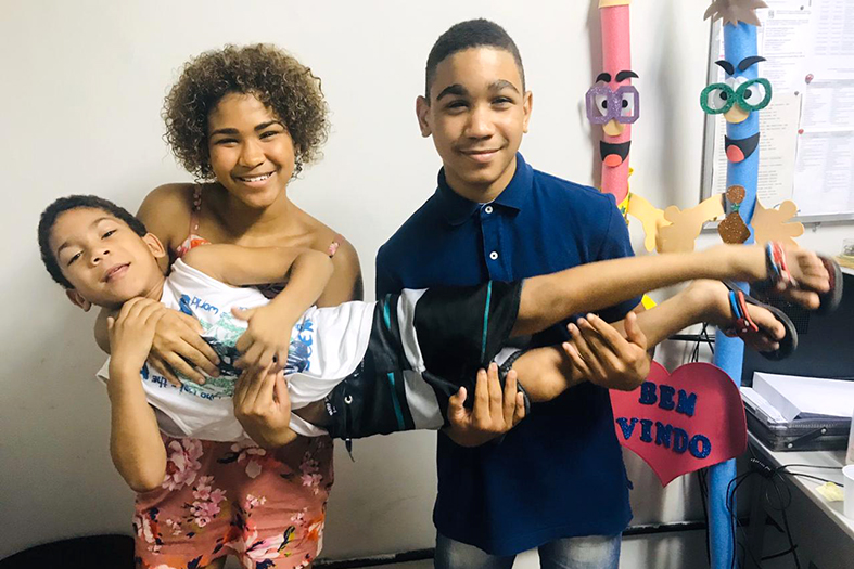 duas crianças, uma menina e um menino, carregam juntos em seus braços uma terceira criança. Ao fundo objeto lúdicos e brinquedos.