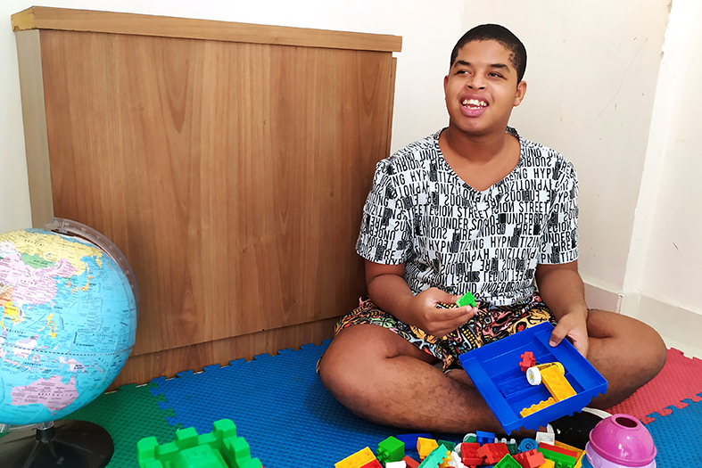 adolescente do sexo masculino de 17 anos, de cor negra, cabelos escuros, usando camisa estampada na cor preta e branca, sentado em piso emborrachado e cercado por brinquedos coloridos