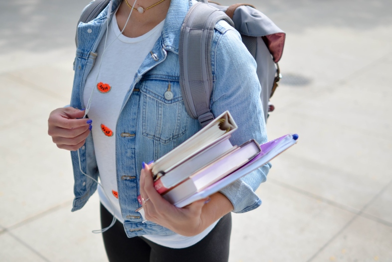 Mulher vestindo jaqueta jeans e segurando livros e cadernos.