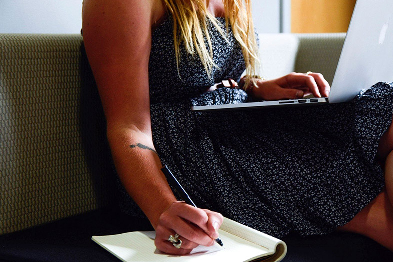 Detalhe de uma jovem estudando com o laptop em seu colo e um caderno de anotações apoiado no sofá.