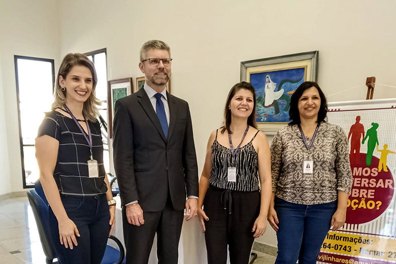 três mulheres e um homem de terno posam para foto com banner temático sobre adoção ao lado.
