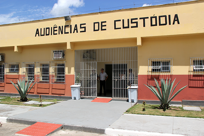 Construção dentro do presídio de Viana. Fachada pintada de amarelo com faixa da cor ocre próxima ao solo. Na parte superior do imóvel, em letras pintadas de preto lê-se "Audiências de Custódia"