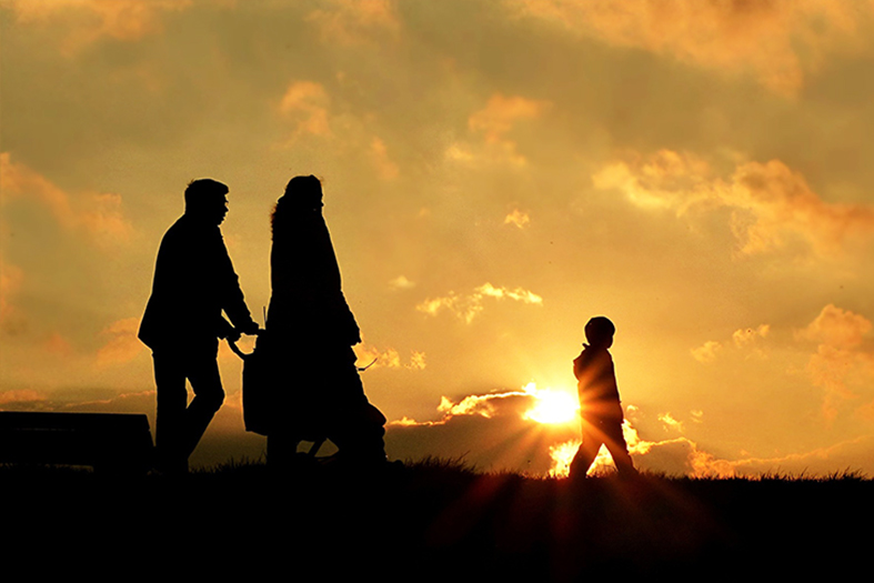 Silhueta de dois adultos, uma criança e um carrinho de bebê, caminhando ao pôr do sol.