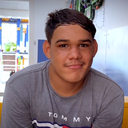 Felipe, adolescente de 14 anos. Ele tem a pele parda, cabelos pretos e olhos castanho-escuros. Ele veste uma camiseta cinza e sorri para a câmera.