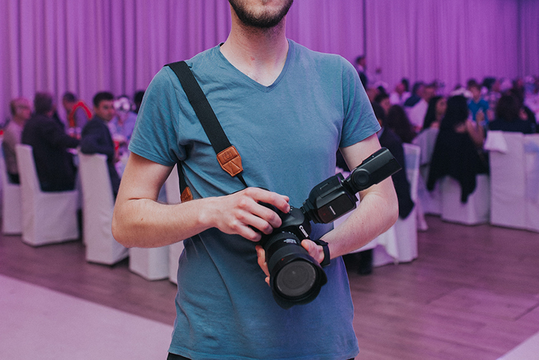Homem carregando uma máquina fotográfica em suas mãos, ao fundo um salão arrumado com a predominância da cor lilás.