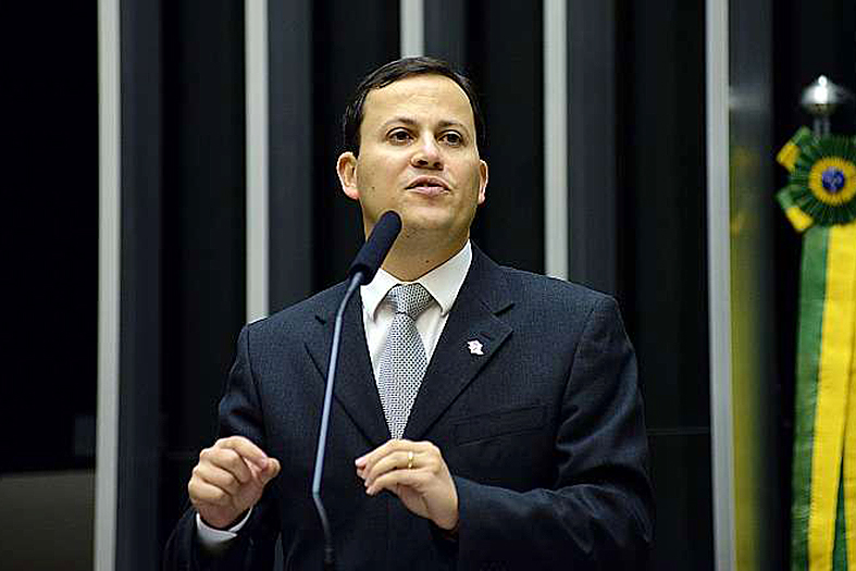 Homem de terno da cor preta, camisa branca e gravata cinza discursa no plenário da câmara dos deputados em brasília. Uma fita verde a amarela está no fundo da imagem à direita.