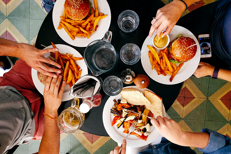 Visão de cima de uma mesa de lanchonete onde quatro pessoas consomem hambúrgueres.