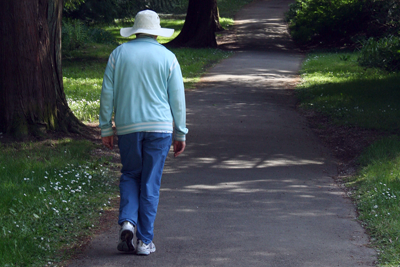 Mulher idosa trajando calça azul escura, camisa de manga comprida de cor azul clara e chapéu branco com largas abas caminha por trecho pavimentado de um parque.