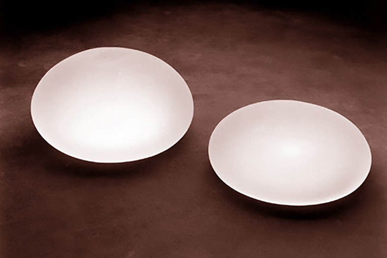 Duas próteses de silicone de cor branca leitosa repousam sobre base da cor marrom.