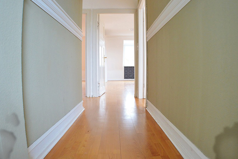 Corredor interno de um apartameento onde se vê em primeiro plano que as paredes de cor creme estão com manchas de infiltração próxima ao rodapé de cor branca.