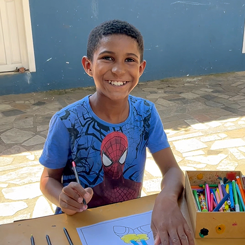 Isaqueu, criança de 10 anos. Ele tem a pele negra e os cabelos encaracolados e de cor preta. Ele está sorrindo para a câmera e na sua frente estão seus desenhos.