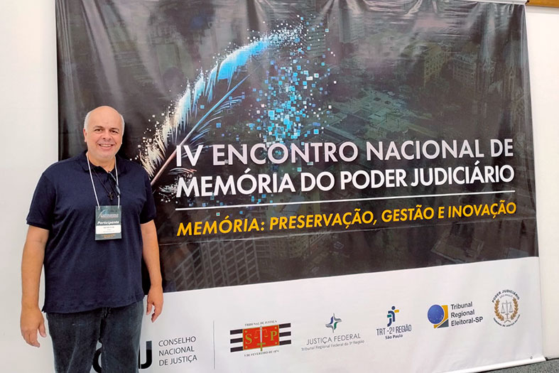 Fábio Buaiz de Lima, coordenador de gestão da informação documental do Tribunal de Justiça do Espírito Santo, posa para foto em frente ao banner do evento "IV Encontro Nacional de Memória do Poder Judiciário".