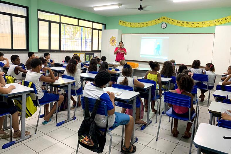 Mulher vestindo camisa rosa fala em uma sala de aula diante de várias crianças.