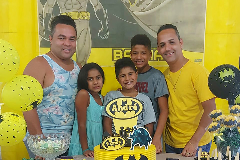 dois homens de estatura mediana e três crianças de estaturas variáveis posam para foto atrás de uma mesa decorada de aniversário