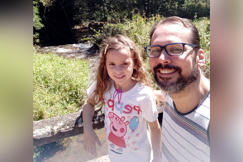 Pai e filha abraçados e sorrindo para a câmera. Eles têm a pele branca. A barba do pai é grisalha, ele é calvo e usa óculos. Os cabelos dela são castanhos claros e são cacheados.