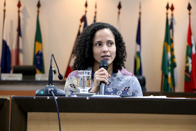 Mulher de pele branca e cabelos da cor preta encaracolados fala ao microfone.