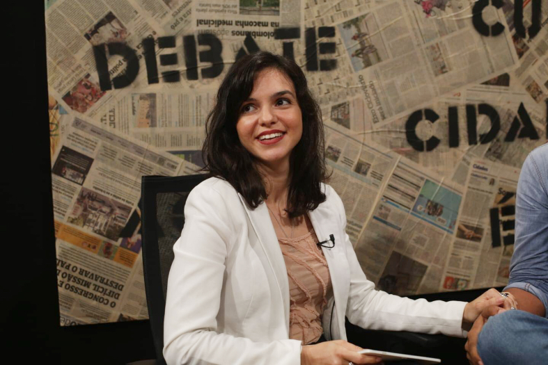 Mulher vestindo casaco branco e camisa bege olha sorrindo para frente.