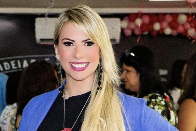 A delegada Cláudia Dematté, Chefe da Divisão Especializada de Atendimento à Mulher. Ela é uma mulher de cor de pele branca, com cabelos tingidos de loiro. Ela veste um blazer na cor azul claro e uma camisa na cor preta.