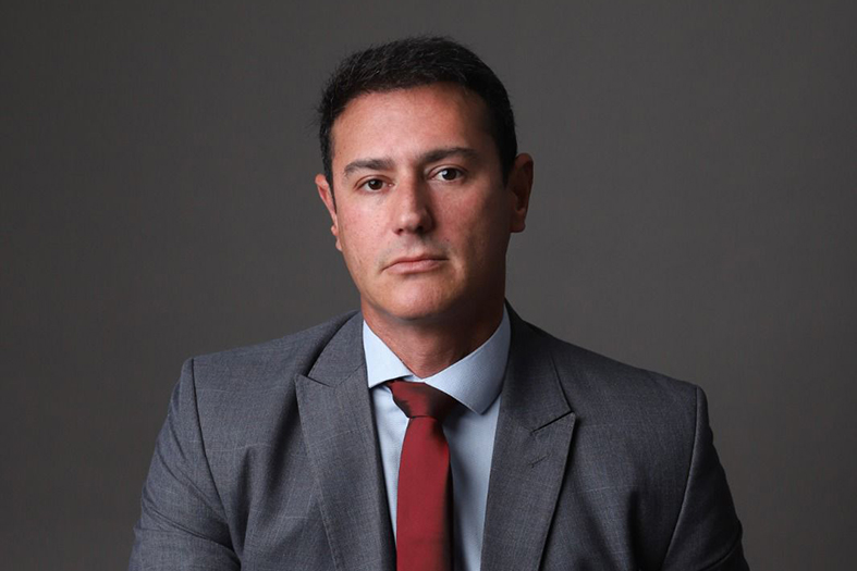 Daniel Blank, homem de pele parda e com cabelos na cor preta, veste um terno na cor cinza e uma gravata vermelha.